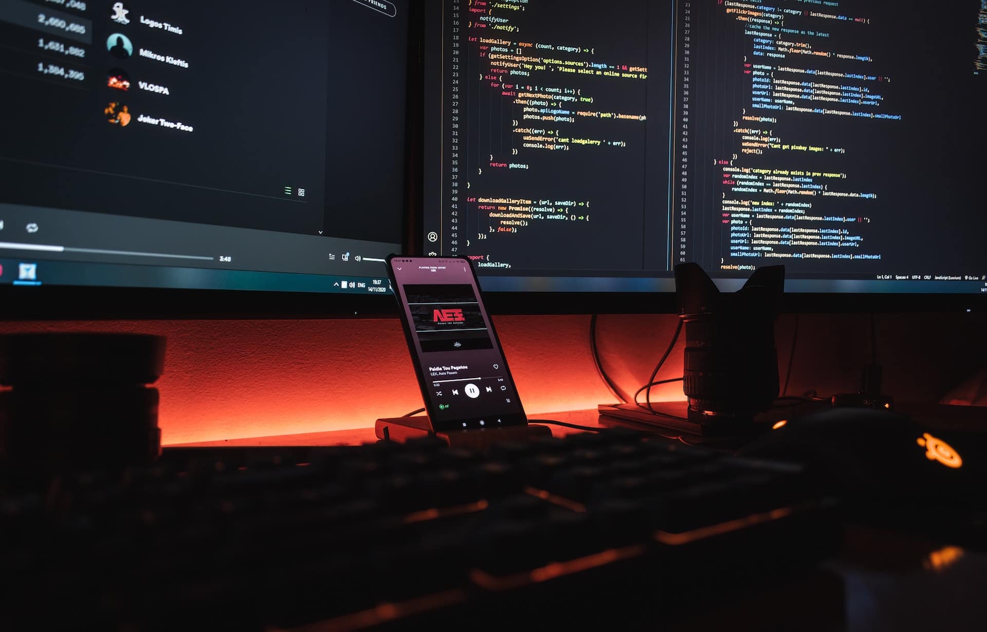 Two monitors with programming code on it. The smartphone is between them with music player on the screen